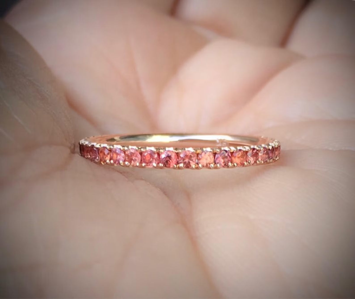 A rose gold eternity ring with a row of orange-small pink gemstones, resting on a person's palm.