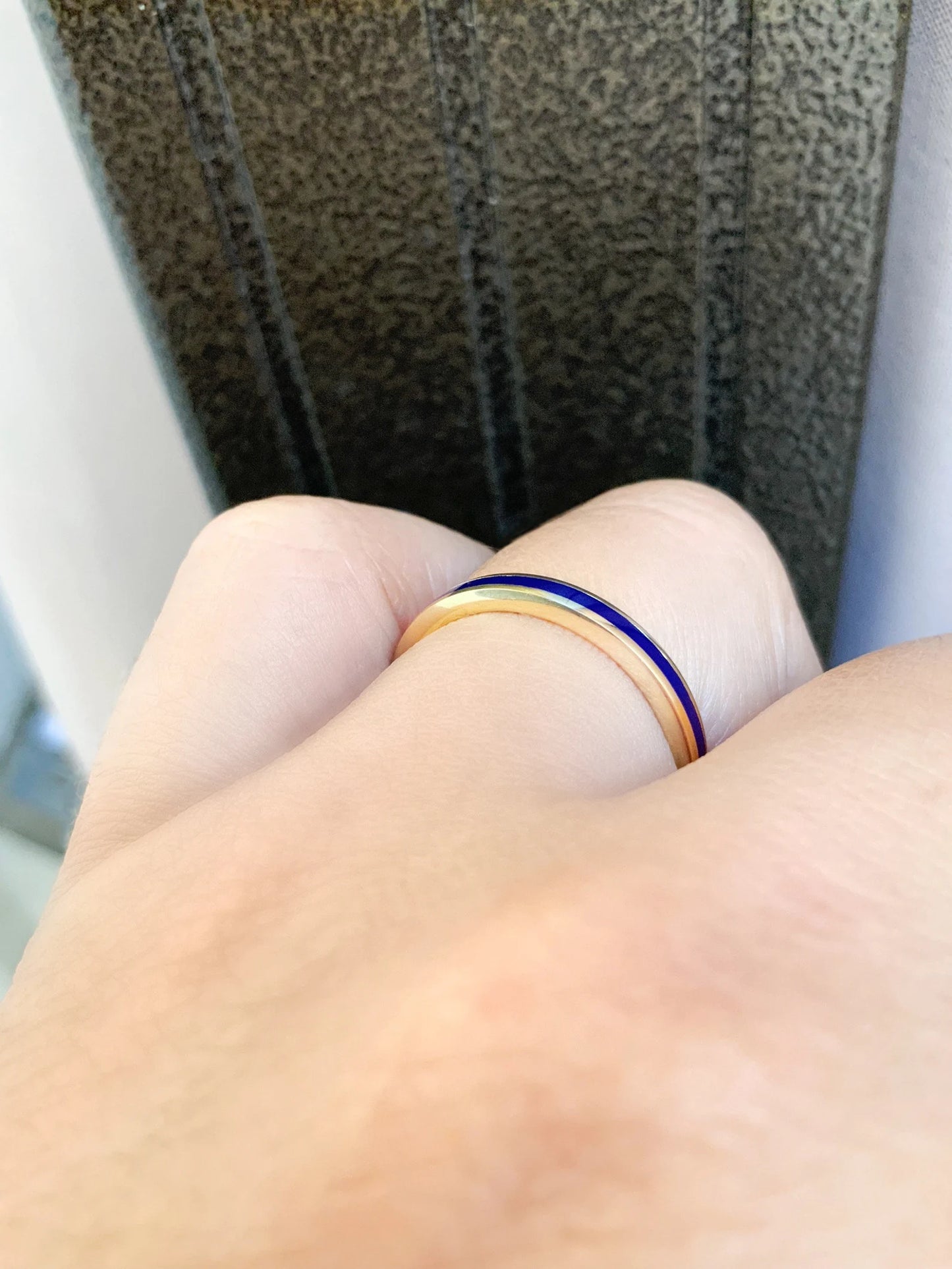 A close-up of a hand wearing two slim rings: one gold band and one dark blue band, set against a textured dark background.