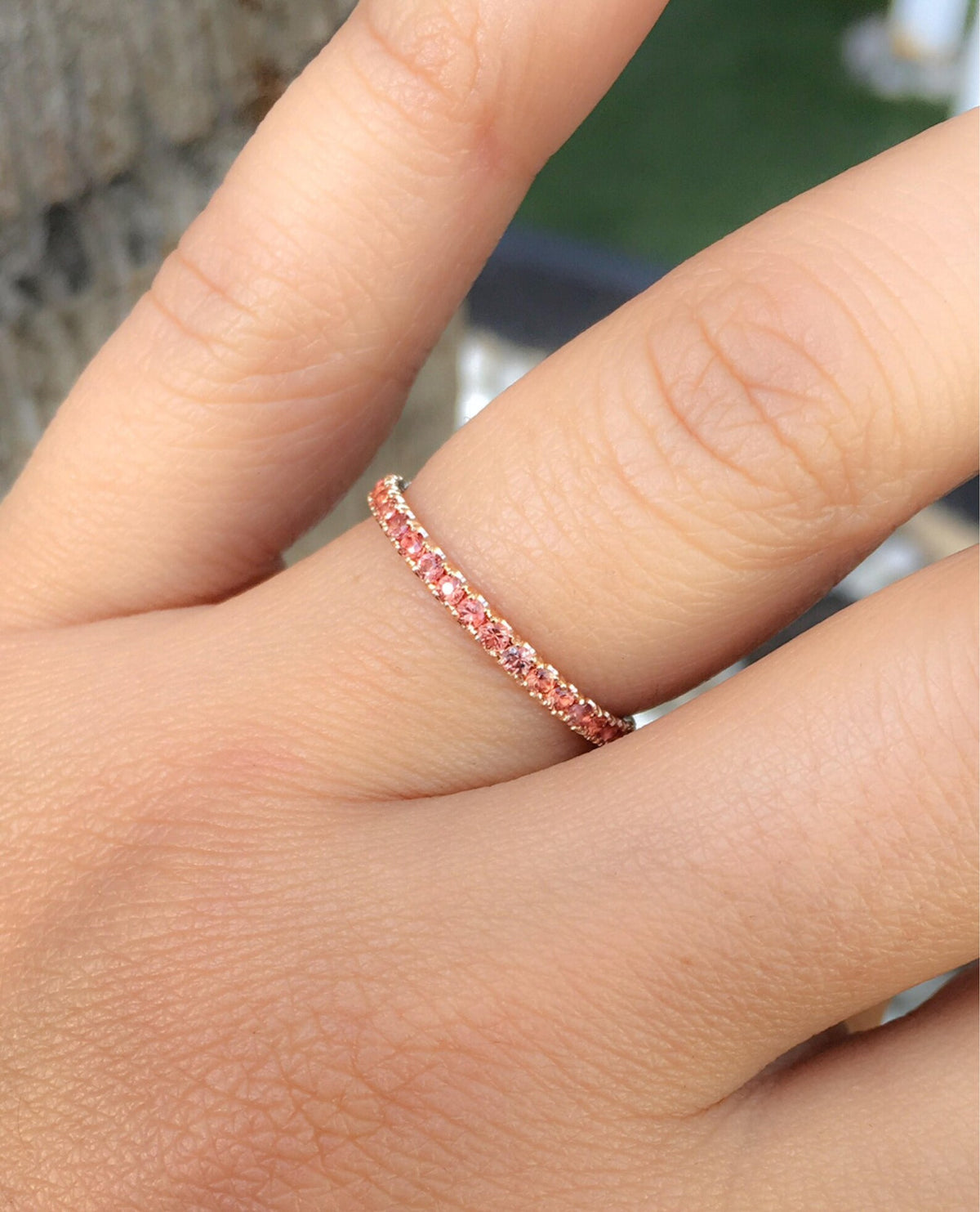 A close-up of a hand wearing a delicate rose gold eternity ring set with small orange-pink gemstones.
