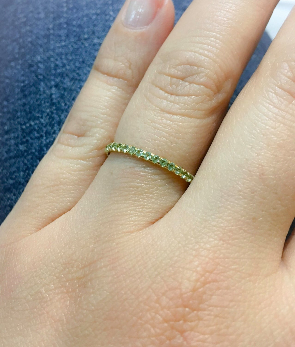 Natural peridot pavé eternity band worn on finger, yellow gold with lime green stones, lifestyle shot against blue background
